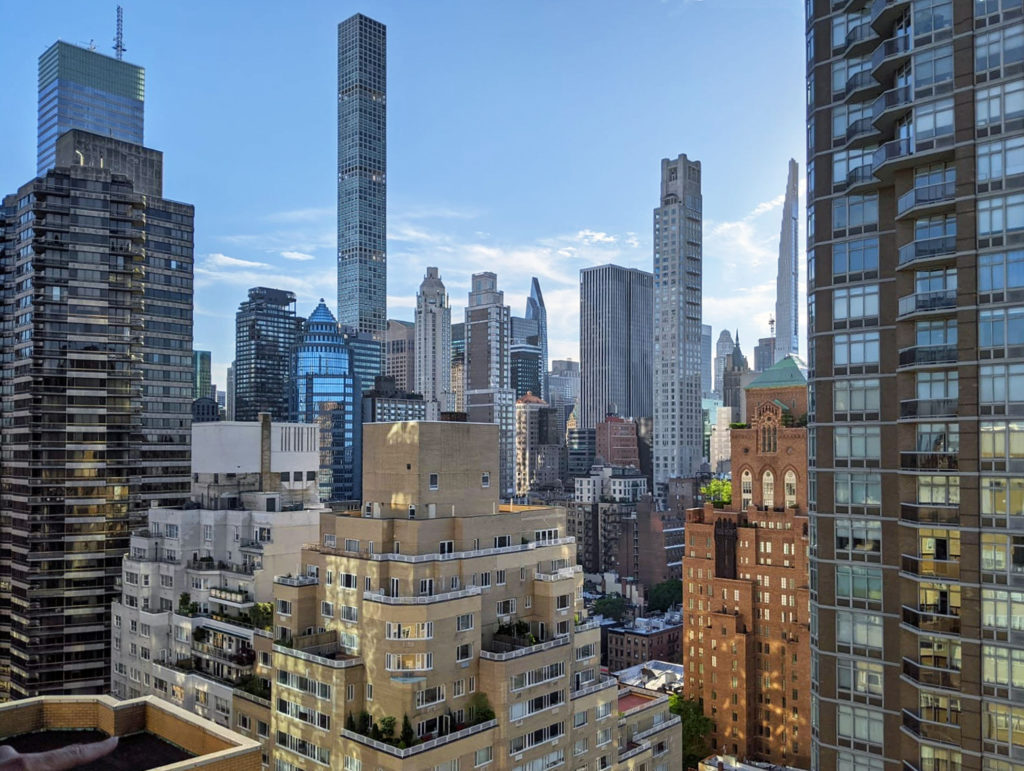 A 13th-story view of equally tall wedding-cake buildings and taller buildings behind, including three so-called pencil towers that are super tall and super thin