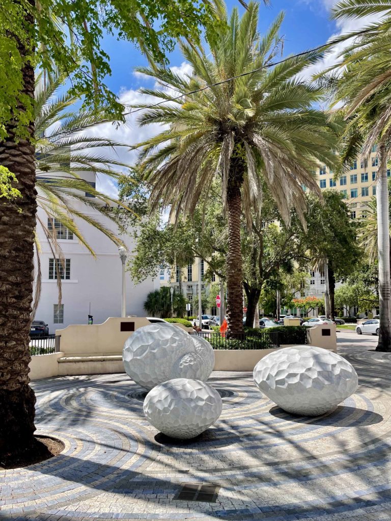Three large stone eggs, each of a different size and uniformly marked with an indented hexagonal pattern are arranged on the pavement of the palm-tree shaded Giralda Plaza in downtown Coral Gables, Florida.