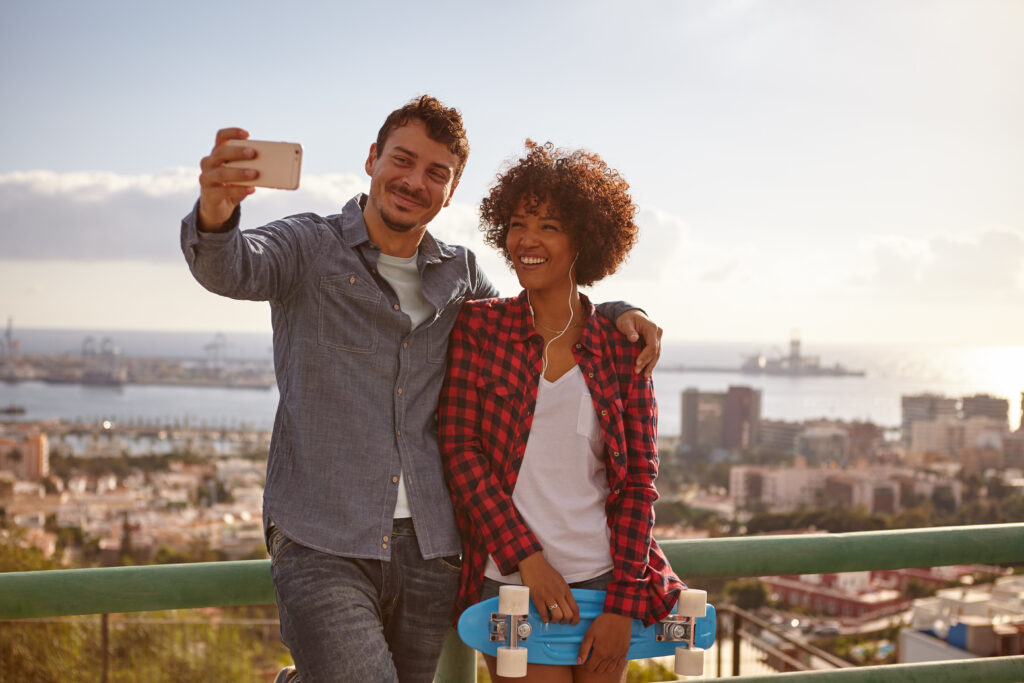 Smiling couple posing with cell phone while she holds a blue skateboard and laughing as they lean against the metal railing of a bridge