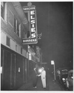 Photograph of two men stand shaking hands on sidewalk underneath Elsie's neon sign, circa 1950s.