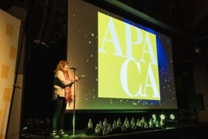 A woman stands on stage smiling at a microphone during an awards ceremony. She holds a piece of paper and is wearing a polka-dotted top, dark pants, and a red scarf. Behind her is a large screen displaying the yellow and white APA California logo, and in front of the stage is a row of glass awards illuminated by green lights.
