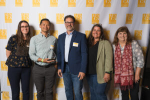Five people pose in front of a backdrop with the APA California Northern Section logo. One person in the middle holds a clear glass award. Everyone is smiling and dressed in business casual attire.