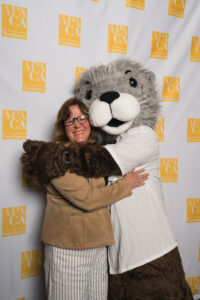 A woman smiles as she hugs a person in a large gray otter mascot costume at the APA California Northern Section event. The background features a step-and-repeat banner with the APA California Northern Section logo.