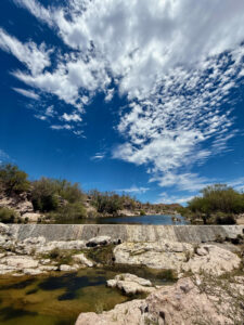 Photograph of Rancho Viejo of Santa Cruz Dam with a beautiful blue sky with puffy white clouds dotting it with rocks ushering a stream of water from the dam.