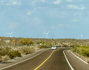 Photograph of a car driving on a two-way highway near Ciudad Constitución, Baja California Sur, Mexico, with wind turbines on a hill rising in the distance.
