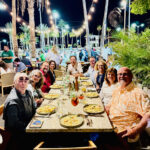 A large group of happy people sitting around an outdoor dining table set for dinner at night with strings of lights hanging above.