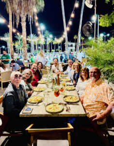 A large group of happy people sitting around an outdoor dining table set for dinner at night with strings of lights hanging above.