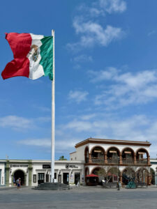 A photograph of the flag of Mexico on a flag pole in front of the Plaza Mijares at the HIstoric Center and Arts District of San Jose del Cabo, Baja California Sur, Mexico.