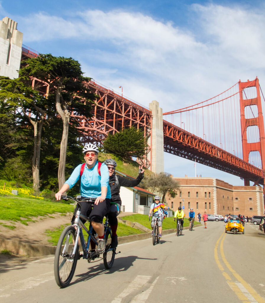 On a road beneath the Golden Gate Bridge, a group of cyclists in single file, led by two women on a tandem, smile as they ride under a partly cloudy sky on a bright day.