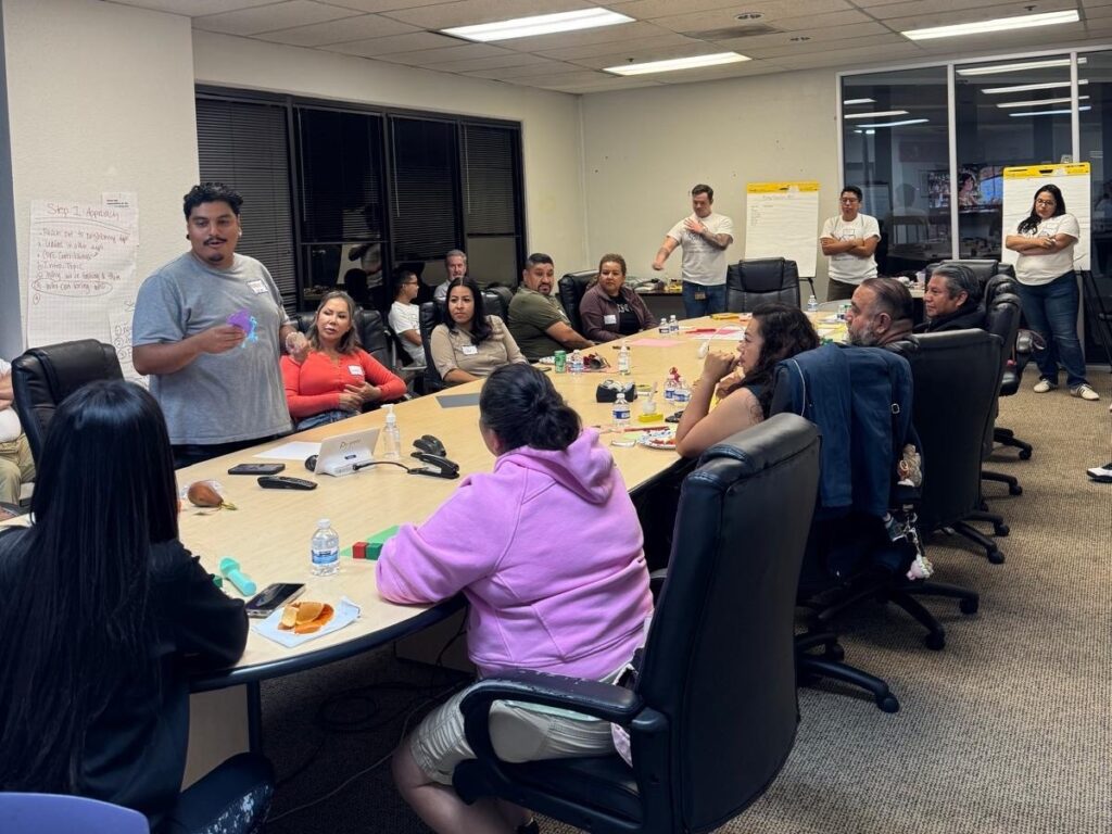 PlaceIt Workshop1_4-3 Image of vendor engagement meeting, discussing the vendor’s find memories of the market. Vendors are sitting around a long, oblong table. Students are standing in the background.