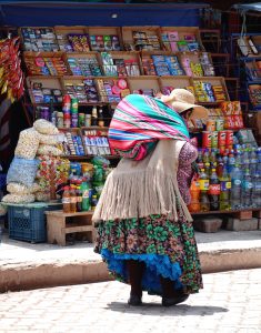 Picture of an indigenous woman carrying a colorful backback walking along the street in front of an outdoor market stand.
