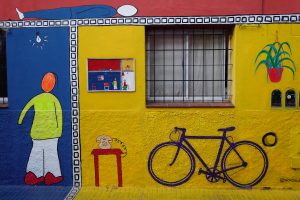 Photograph of the outside facade of a home in Buenos Aires, Argentina, with yellow wall, characture of person walking into a blue door to the left with a picture, table and wall hangings painted under and around the window.
