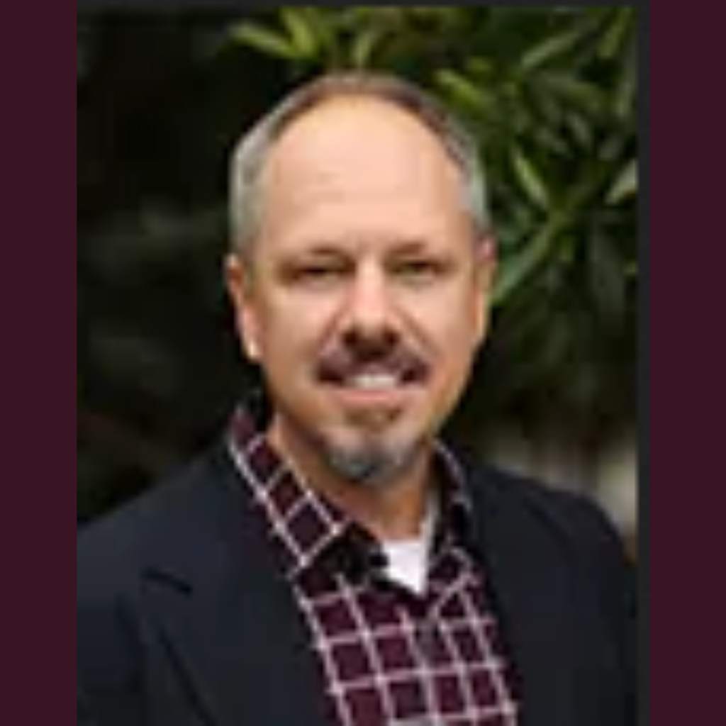 Headshot of a smiling middle-aged Caucasian man with graying hair, a mustache, and a goatee, wearing a maroon plaid shirt with a black sweater.