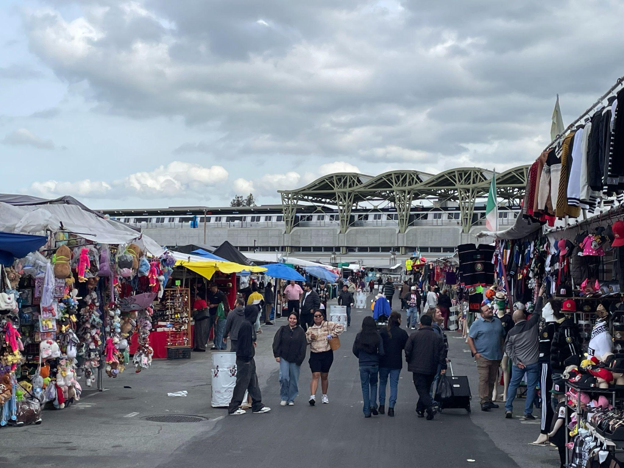 Berryessa & BART A crowded outdoor marketplace with colorful vendor tents and shoppers, featuring the elevated Berryessa BART station and a train in the background.